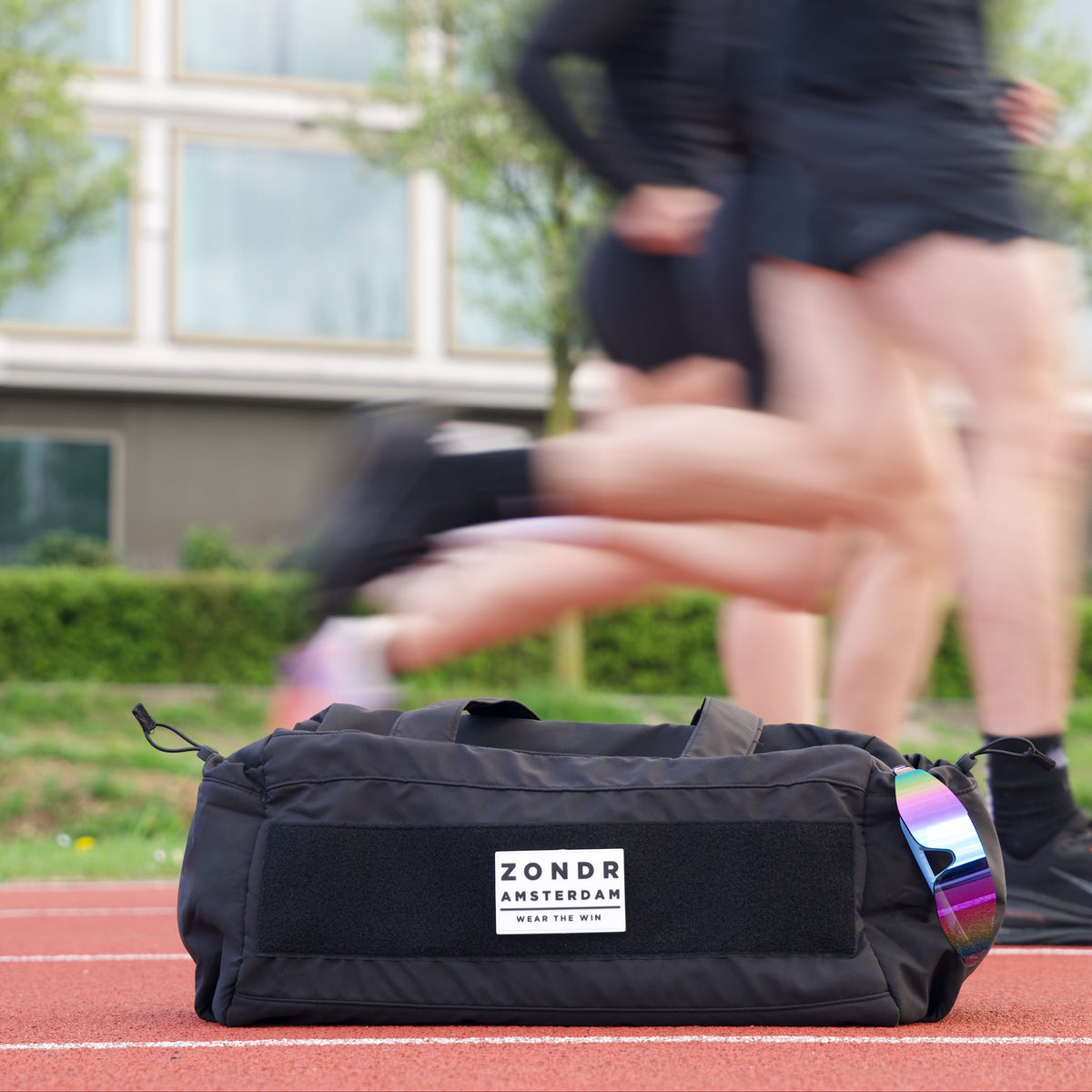Black ZONDR WEAR THE WIN duffel bag on a running track with athletes training behind it, iridescent sunglasses tucked into the end pocket