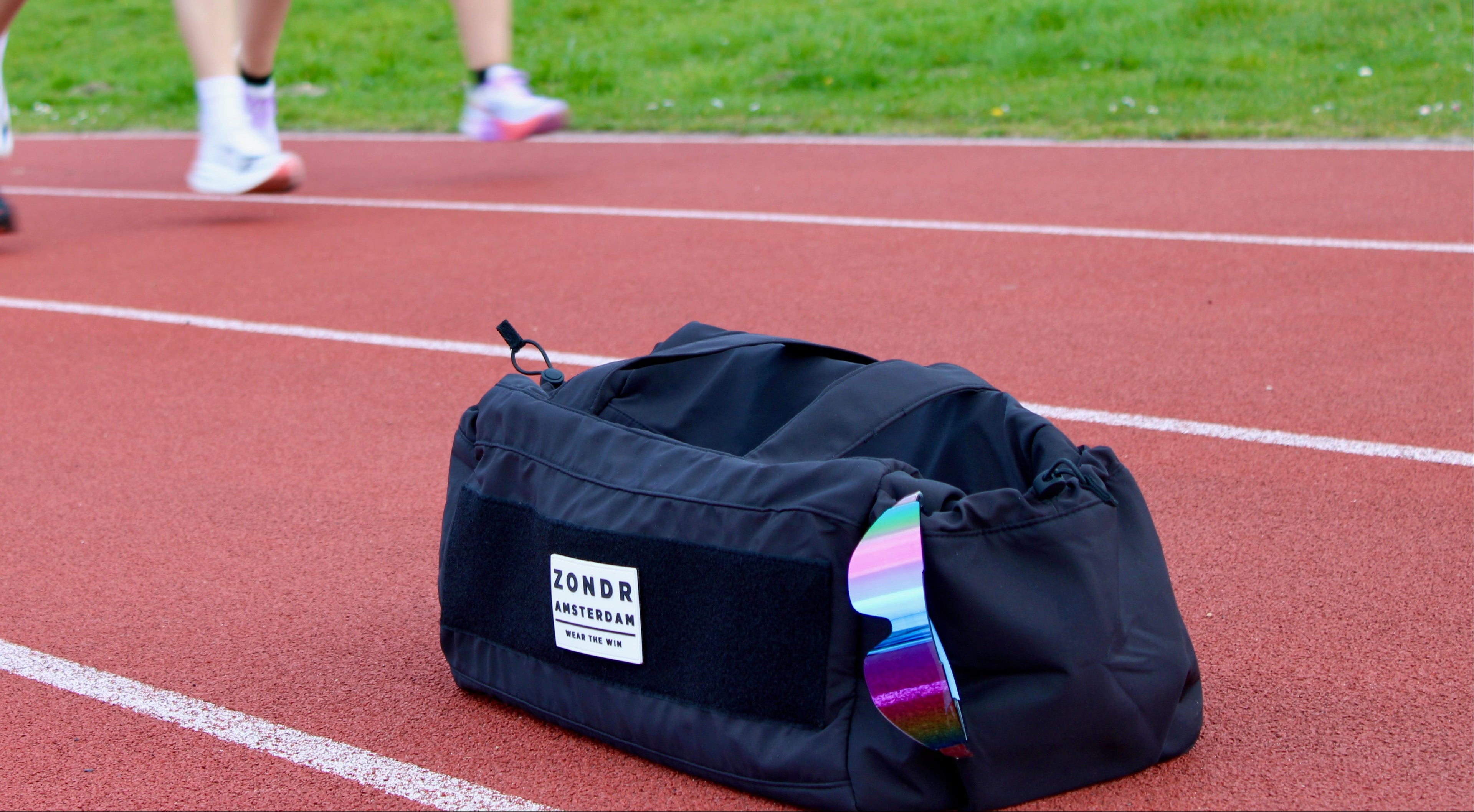 the black zondr duffel bag sits on a red running track in the foreground, with blurred runners on the track in the background.