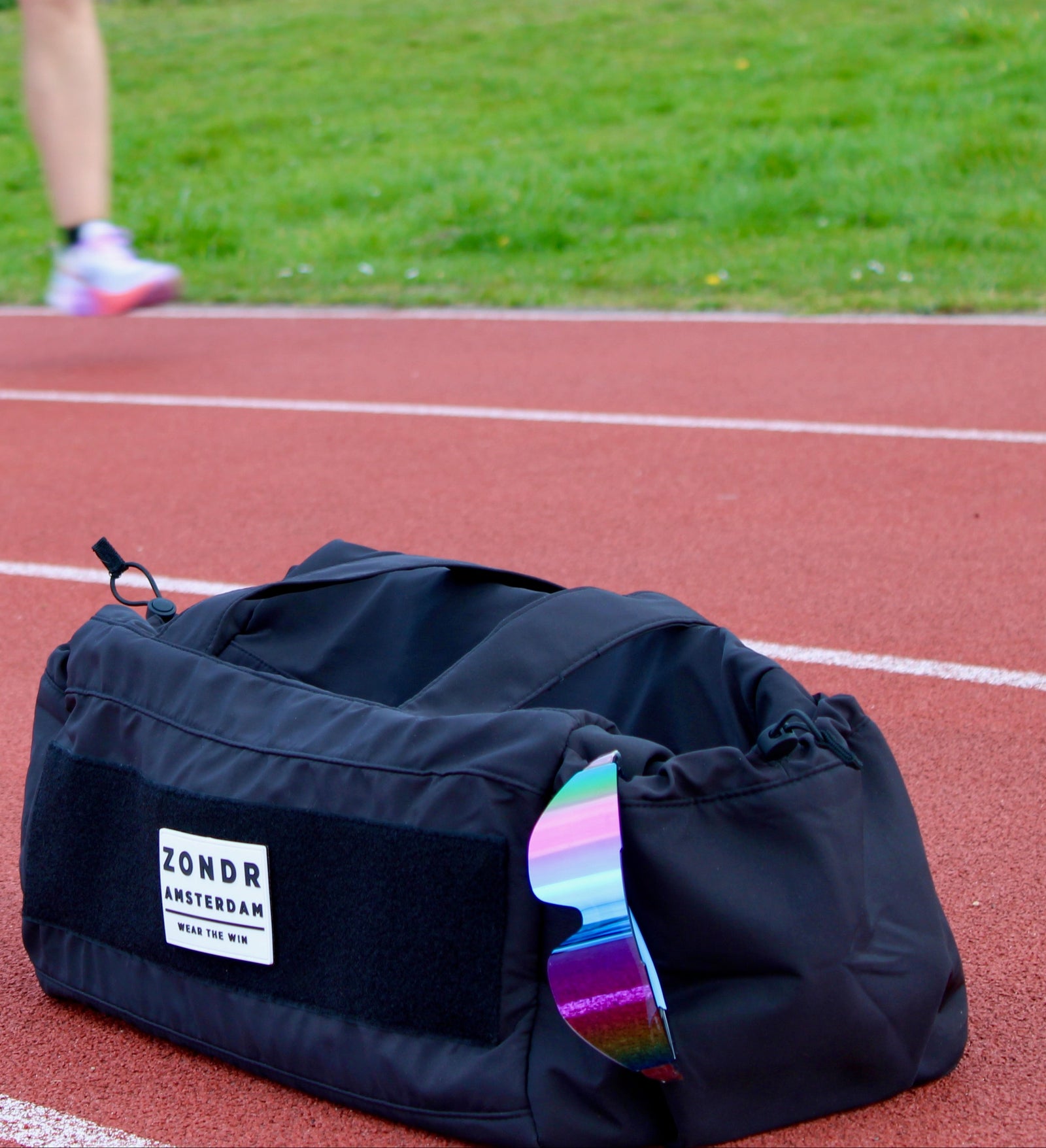 the black zondr duffel bag sits on a red running track in the foreground, with blurred runners on the track in the background.